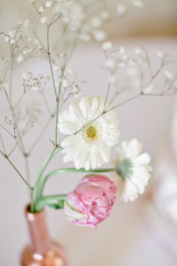 Close-up detail shot of a small floral arrangement in a copper or rose gold bud vase. The arrangement includes a white gerbera daisy, a smaller white daisy, a soft pink ranunculus, and sprigs of white gypsophila (baby's breath). The overall color palette is white, blush pink, and soft green against a pale, blurred neutral background. The styling is delicate and minimal, consistent with a romantic or soft classic aesthetic. The shallow depth of field keeps the focus on the central white gerbera while the background remains softly out of focus.