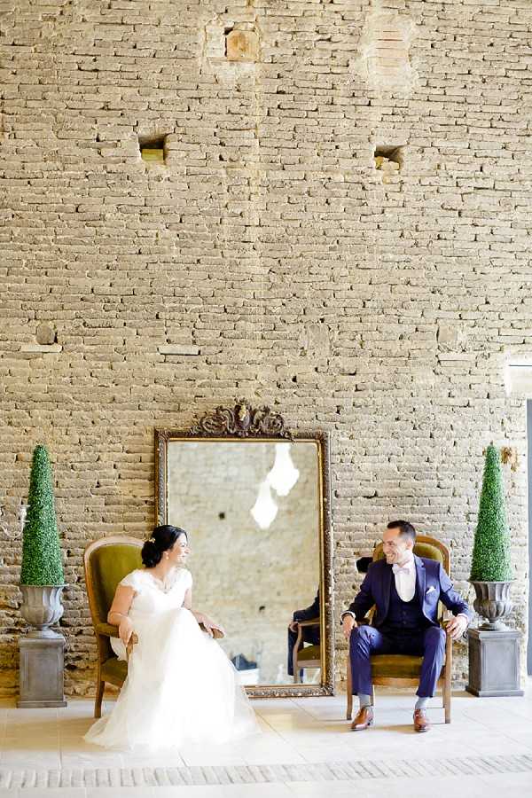 A couple portrait taken indoors against a tall exposed brick wall with a rustic, aged texture. The bride is seated in a vintage wooden armchair on the left, wearing a white cap-sleeve lace gown with a full tulle skirt and floral hair accessories, while the groom sits in a matching chair on the right in a navy three-piece suit with a light pink tie and brown shoes. Between them is a large ornate gold-framed floor mirror, flanked by two cone-shaped boxwood topiaries in grey stone urns. The styling combines classic French château decor with rustic architectural elements. Wide portrait shot capturing the full setting.