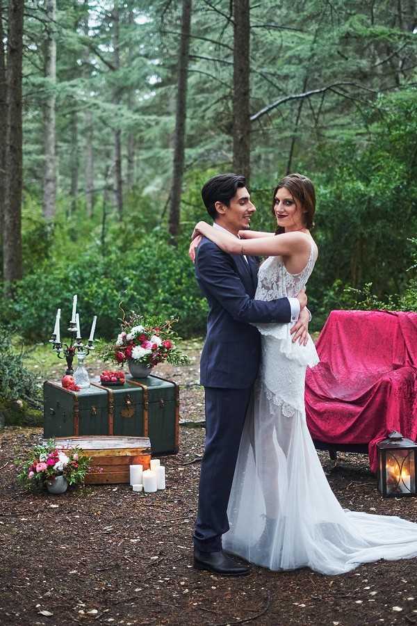 A couple poses together in an outdoor forest setting, embracing and facing each other in a portrait-style shot. The bride wears a fitted white lace gown with a low back, sheer overlay, and a small train, while the groom wears a navy blue suit. Behind them, a styled vignette includes a dark green vintage trunk topped with a black candelabra with white taper candles and a floral arrangement of deep red and pink blooms, alongside a second open wooden trunk at ground level with white pillar candles and a low arrangement of pink and red flowers. To the right, a sofa draped in a deep crimson/magenta velvet throw and a lit metal lantern on the ground complete the scene. The overall styling theme is moody and romantic with a dark jewel-tone palette, mixing vintage travel trunks, candlelight, and rich red florals in a pine forest clearing.