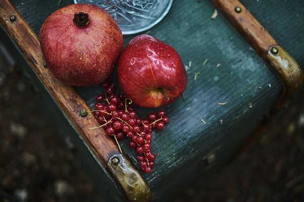 A close-up detail shot of a styled flat lay arrangement on a weathered teal-painted wooden chair, featuring a deep red pomegranate, a red apple with water droplets, and a scattered cluster of red currants on stems. A cut crystal glass is partially visible in the upper background. The overall color palette is deep red and burgundy against the distressed teal wood, suggesting an autumn or harvest-themed wedding styling. The image is slightly moody and dark in tone, consistent with an editorial autumn wedding aesthetic.