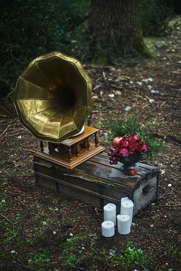 A styled wedding decor detail shot set outdoors in a wooded area. A brass gramophone sits on a small ornate wooden stand atop a rustic weathered wooden trunk, alongside a dark metal compote vase filled with deep crimson ranunculus, dark red berries, pomegranate, and dark green foliage. Four white pillar candles of varying heights are placed on the ground in front of the trunk. The overall styling palette is dark and moody — deep reds, gold, and dark wood tones — consistent with a romantic, vintage-rustic or dark forest wedding aesthetic. Close-up detail/still life composition.