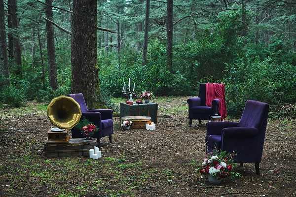 An outdoor styled wedding lounge vignette set within a dense forest, with no people present. Three deep navy-purple velvet armchairs are arranged around a dark green vintage trunk used as a coffee table, on which white taper candles in black candlesticks and a small floral arrangement of red and white blooms are placed. A brass gramophone horn sits on a stack of wooden crates to the left, accompanied by small white pillar candles on the ground. A red draped fabric hangs from a chair on the right side, and a loose floral arrangement of white and red flowers sits on the forest floor in the foreground. The overall styling theme is dark, moody, and vintage-romantic with a jewel-toned color palette of deep purple, crimson, and gold. Wide shot taken at ground level.