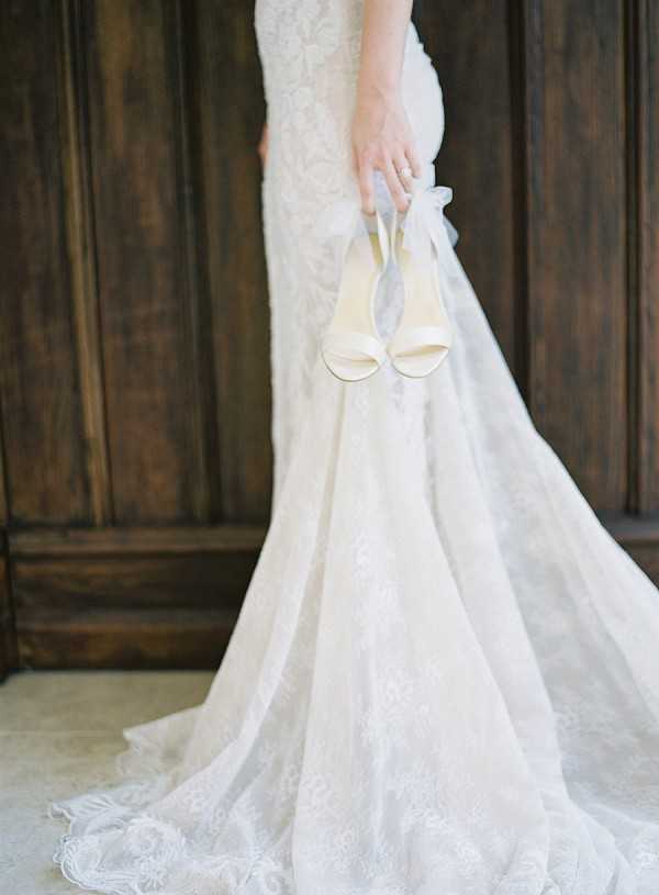 A detail portrait of a bride holding a pair of ivory satin open-toe heeled sandals with ribbon ankle ties, shot from the torso down against a dark wood paneled door interior. The bride is wearing a fitted lace gown with a floral lace pattern and a long cathedral-length train that pools on the tiled floor. A diamond ring is visible on her hand. The image has a soft, film-style tone with warm, diffused light. Close-up portrait composition emphasizing the dress silhouette and shoes as intentional styling details.