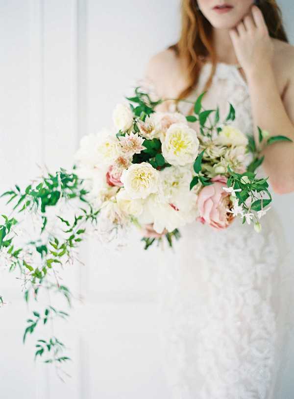A close-up portrait of a bride holding a loose, garden-style bridal bouquet composed of cream and blush garden roses, white dahlias, soft pink peonies, small blush astrantia, and trailing green foliage with delicate white jasmine-like blooms cascading to one side. The bride is wearing a sleeveless ivory lace gown with a halter neckline, and her face is partially cropped at the top with one hand raised near her chin. The background is a soft, bright white and appears to be an interior wall, giving the image a clean, airy, fine-art film aesthetic. The composition is a medium close-up focused on the bouquet, with the bride's torso and lower face softly out of focus.