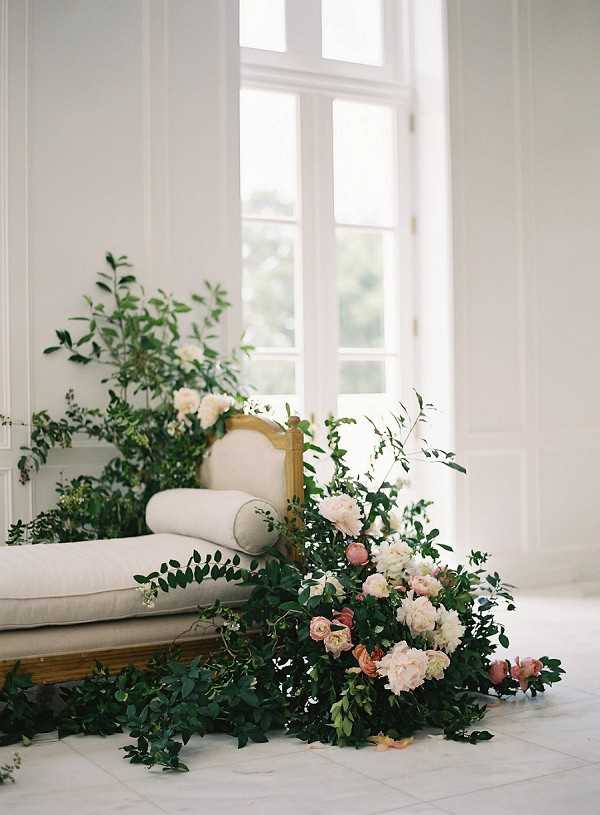 A close-up detail shot of a floral installation styled around a French-style cream upholstered daybed or settee with a gilt wood frame, set inside a bright white-walled interior room with tall panelled French doors. A large, loosely arranged floor-level floral installation spills from the base and side of the settee across a pale stone-tiled floor, featuring blush and cream peonies, dusty pink garden roses, and abundant dark green foliage including eucalyptus and mixed trailing greenery. Additional greenery and white blooms are arranged behind the settee near the window. The overall styling is romantic and organic with a garden-gathered aesthetic, using a soft blush, cream, and deep green palette against the all-white interior. No people are present in the image.
