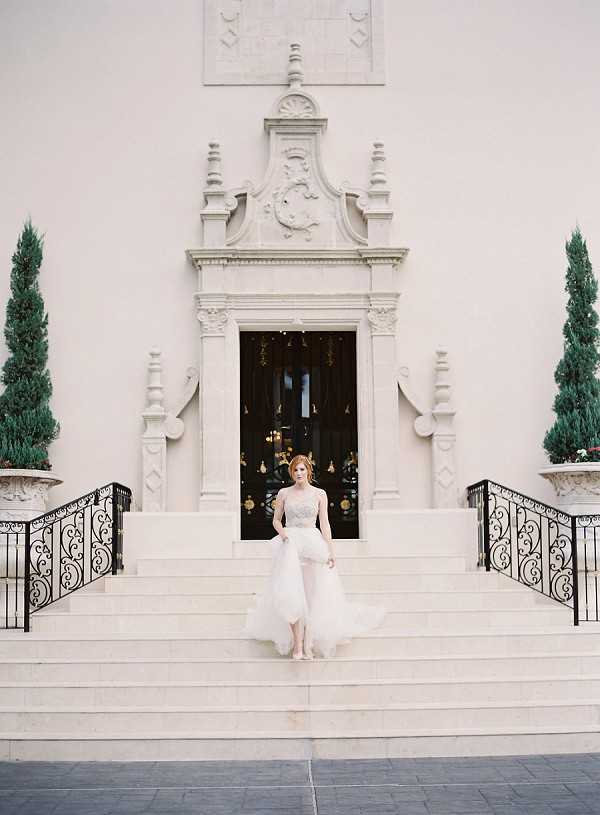 A bridal portrait shot outdoors on the grand entrance steps of a large formal building featuring ornate white stone architectural detailing, an elaborately carved doorway pediment, and flanking topiary cypress trees in classical urns. The bride, a redhead, stands center-frame on the steps wearing a strapless ivory gown with a beaded bodice and a layered tulle skirt that appears to be in motion. Black wrought iron scrollwork railings frame either side of the wide staircase. The composition is a full-length portrait taken straight-on with the architectural facade as a backdrop, giving the image a classic, formal feel. Potential venue feature image.
