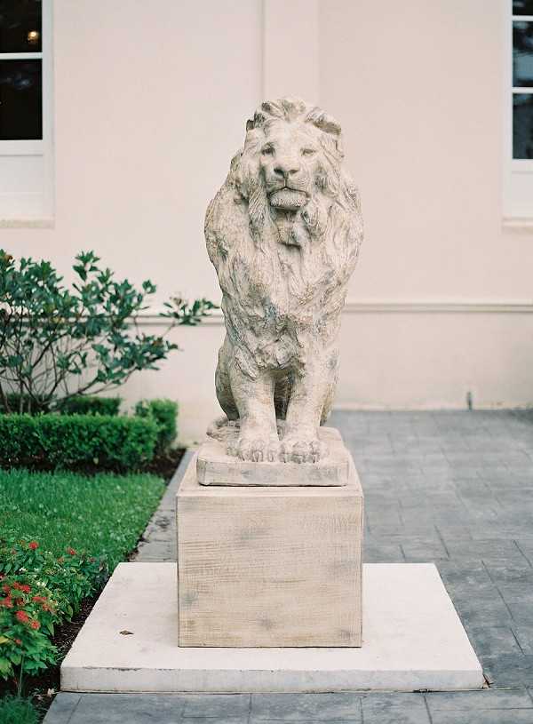 A close-up exterior shot of a large stone lion statue mounted on a square stone pedestal, positioned on a paved courtyard area in front of a cream-rendered building with white-framed windows. The statue is weathered with aged patina and is flanked by manicured boxwood hedges, green lawn, and small red flowering shrubs. No people are present in the frame. The image has a clean, architectural quality typical of venue detail photography shot on film. Potential venue feature image.