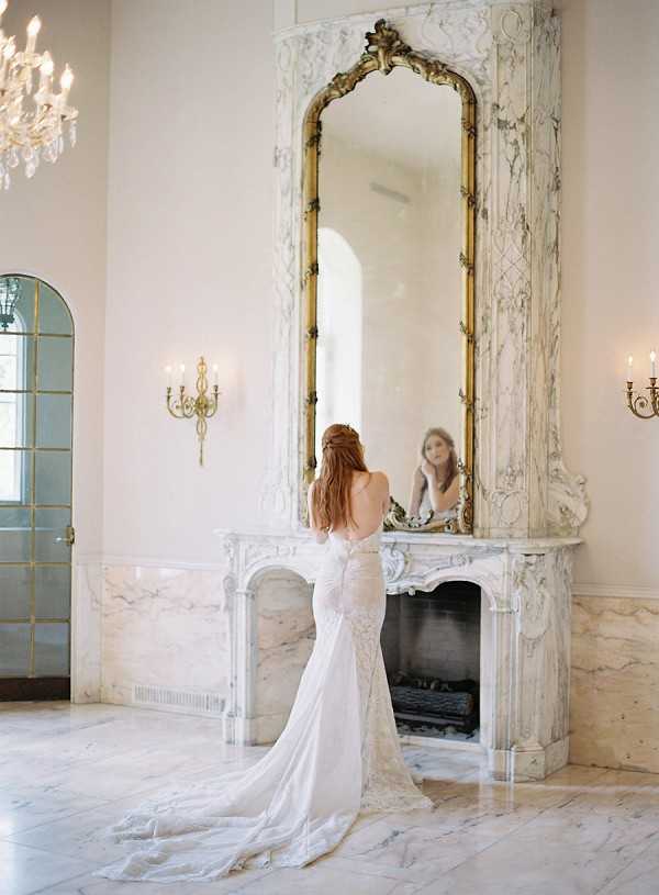 A bride stands alone in front of an ornate marble fireplace inside a grand interior room, viewed from behind as she examines her appearance in a tall floor-to-ceiling mirror with a gilded gold baroque frame. She wears a fitted ivory lace wedding gown with an open back, a low-cut back detail, and a trailing lace train pooling on the marble floor. Her auburn hair is styled in a loose half-up arrangement. The room features cream and grey veined marble on the fireplace surround and floors, gold candle wall sconces, a crystal chandelier visible in the upper left corner, and an arched French door with gold trim to the left. The portrait-style composition captures both the bride's back and her reflection simultaneously, emphasizing the classic French interior architecture. Potential venue feature image.