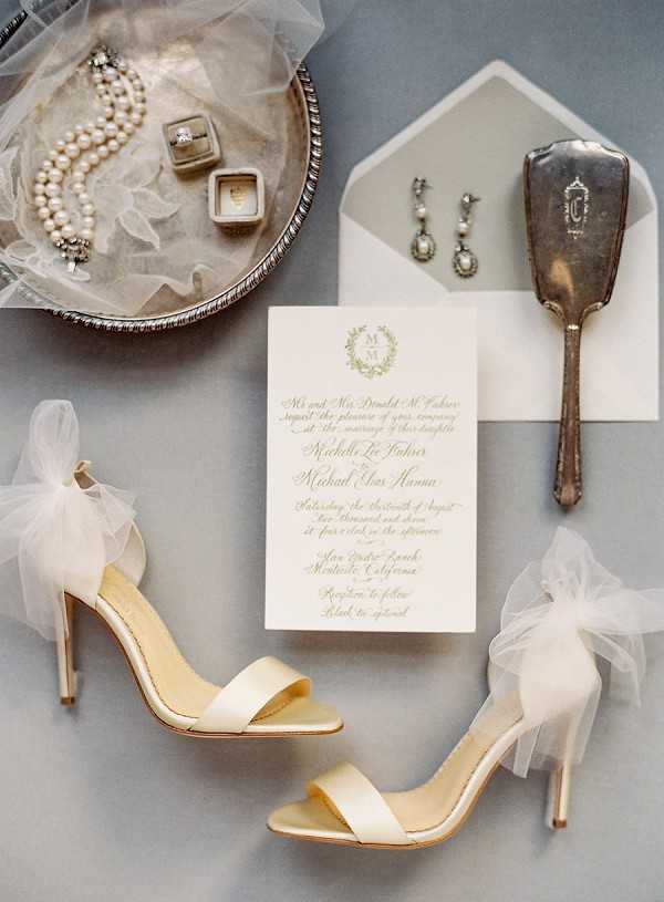 A flat lay detail shot of bridal accessories arranged on a soft grey surface. The composition includes a pair of ivory satin stiletto heeled sandals with tulle bow embellishments at the ankle, a formal wedding invitation printed in sage green calligraphy script with a monogram wreath crest, a grey envelope liner, drop earrings featuring pearls and crystals, an antique silver hand mirror with monogram engraving, a pearl bracelet, and two ring boxes — one containing a diamond engagement ring — displayed in a silver tray with white tulle. The overall styling palette is ivory, soft gold, and grey with classic, formal detailing.