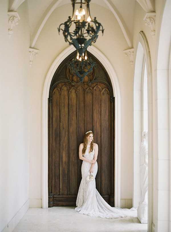 A bridal portrait taken indoors at what appears to be a château or historic building, with the bride standing in front of a tall, ornate arched wooden door with Gothic-style carved paneling. The bride wears a fitted, lace mermaid-style gown with a trailing hem and holds her shoes in one hand, looking upward with a relaxed expression. She has long auburn hair and wears a delicate floral crown or tiara. An ornate wrought-iron chandelier with candle-style lights hangs above, and white arched stonework frames the composition on both sides. The overall styling is classic and refined, with a muted, soft palette. Full-length portrait shot, centered composition. Potential venue feature image.