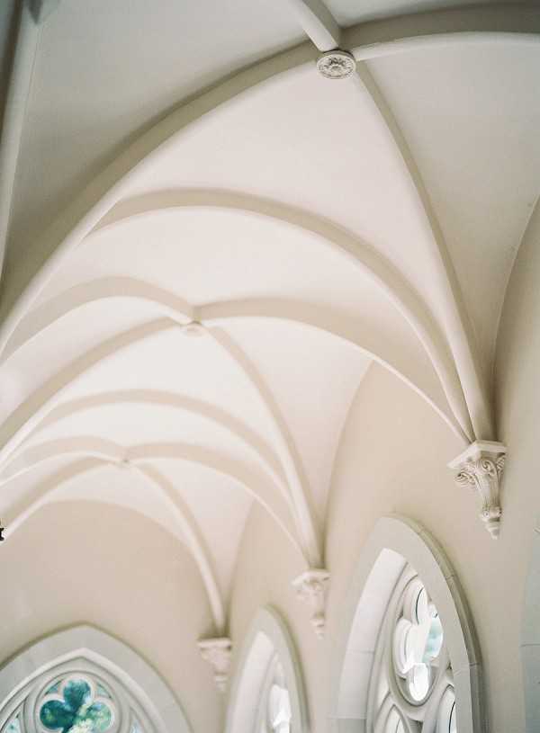 A close-up architectural detail shot of the interior of a chapel or church, showing a series of white Gothic ribbed vaulted ceilings receding into the background. The arched ribs meet at carved corbels along the right wall, and a row of Gothic arched stained glass windows with teal and pale blue patterning is visible along the lower edge of the frame. No people are present in the image. The composition is an upward-angled detail shot emphasizing the repetition of the arch forms and the soft white-on-white tonal contrast of the plasterwork. Potential venue feature image.