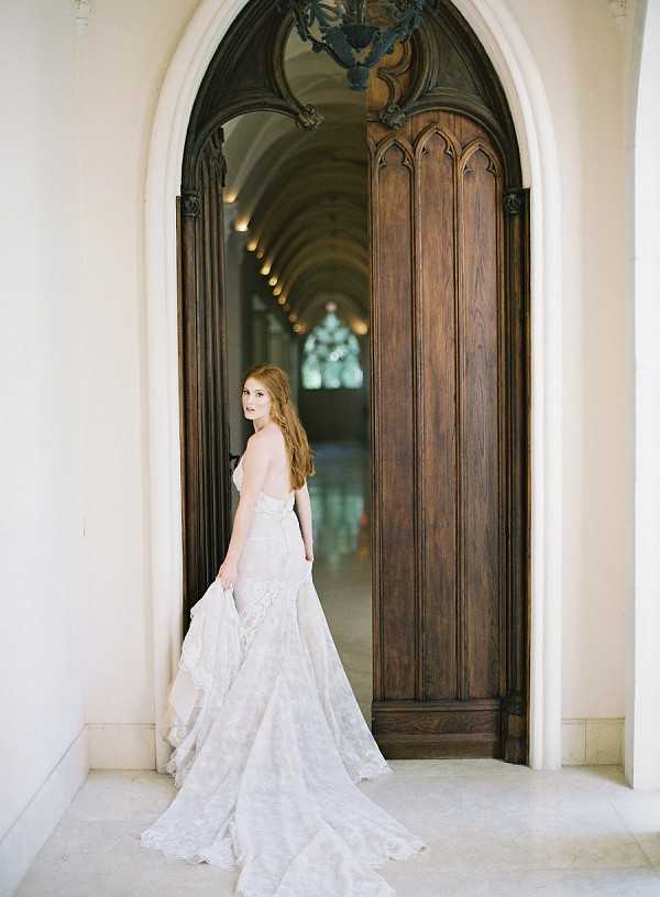 A bridal portrait of a single woman with long auburn hair, wearing a strapless lace wedding gown in an ivory-champagne tone with a full train that pools on the marble floor. She stands in the open doorway of a grand arched entrance with ornate dark wood Gothic-style doors, glancing back over her shoulder toward the camera. Behind her, a vaulted corridor with repeating arched ceilings and a chandelier recedes into the distance, suggesting a chateau or historic manor interior. The shot is a full-length portrait with a classic, editorial styling approach that emphasizes the architectural details of the venue alongside the gown's lace texture and dramatic train. Potential venue feature image.