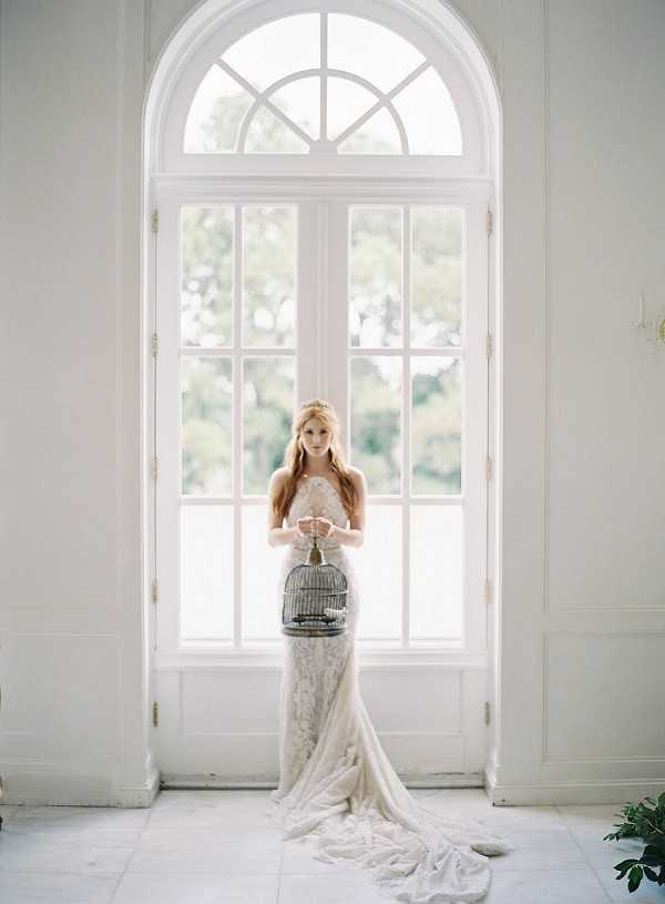 A bridal portrait shot indoors, with a bride standing centered in front of a tall arched window with white painted woodwork and multi-pane glass. The bride wears a strapless ivory lace fitted gown with a long trailing skirt pooling on the marble tile floor, and her long auburn hair falls loosely over her shoulders with a delicate gold hair accessory. She holds a small ornate metal birdcage in front of her at waist height, styled as a decorative prop. The overall aesthetic is romantic and editorial, with a classic French interior setting featuring white walls, tall architectural windows, and pale stone flooring. The image is a full-length portrait composition with soft, diffused natural light flooding in from the window behind her.