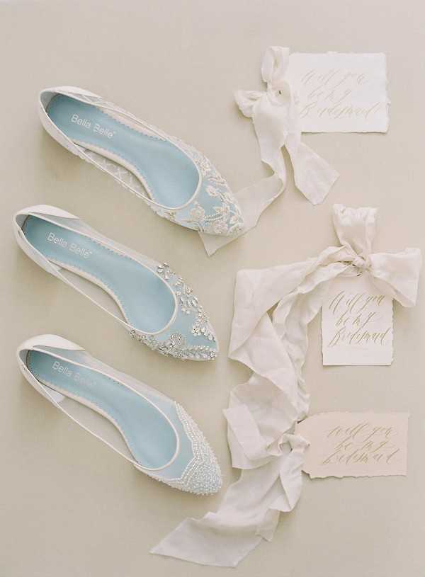 A flat lay detail shot featuring three pairs of Bella Belle bridal ballet flats arranged vertically on a neutral cream background. Each shoe has a powder blue insole and white embellished upper in different styles: the top pair features delicate lace appliqué, the middle pair has crystal and rhinestone floral embellishments, and the bottom pair is covered in pearl beading. To the right of each pair of shoes is a handwritten calligraphy card reading 'Will you be my bridesmaid?' on cream deckle-edged paper, each tied with a flowing ivory silk ribbon. The overall styling palette is white, cream, and powder blue with a classic, refined aesthetic.