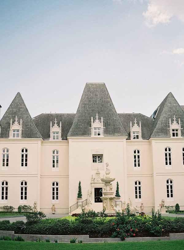 Wide exterior shot of a French chateau-style venue featuring cream-rendered walls, multiple pointed slate-grey turret roofs with decorative Gothic-style dormers, and tall arched white-framed windows. A ornate stone tiered fountain serves as the central focal point in the forecourt, flanked by paired Italian cypress trees and low sculpted hedging. The formal entrance features stone steps with iron railings and symmetrical garden beds. No people are visible in the frame. Potential venue feature image.