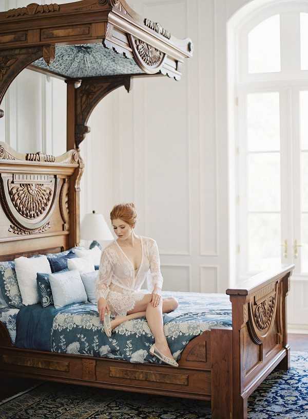 A bridal getting-ready scene shot indoors in a bedroom with white paneled walls and large arched windows. The bride, a red-haired woman with her hair pinned up, sits on an ornately carved dark walnut four-poster canopy bed, wearing a sheer white lace robe and putting on embellished flat bridal shoes. The bed is dressed in deep teal and white floral printed linens with an assortment of blue and white throw pillows. A blue and white patterned area rug lies on the hardwood floor. The overall decor palette combines rich dark wood tones with teal and white, giving the room a classic, period-inspired aesthetic. Medium-distance portrait shot with the architectural bed frame as a dominant compositional element.