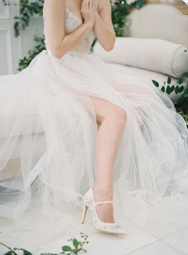 A close-up detail portrait of a bride seated on a cream upholstered sofa during a getting-ready or styled shoot moment, indoors. The frame captures her torso and legs, highlighting her ivory tulle ballgown skirt with a lace beaded bodice, along with ivory pointed-toe mary-jane stiletto heels featuring a delicate floral or lace overlay pattern. The skirt billows across the surface in soft, layered tulle. Green foliage with small white berries is arranged around the sofa as decorative styling. The overall palette is ivory and white with touches of green, and the aesthetic is romantic and soft with a fine art film quality.