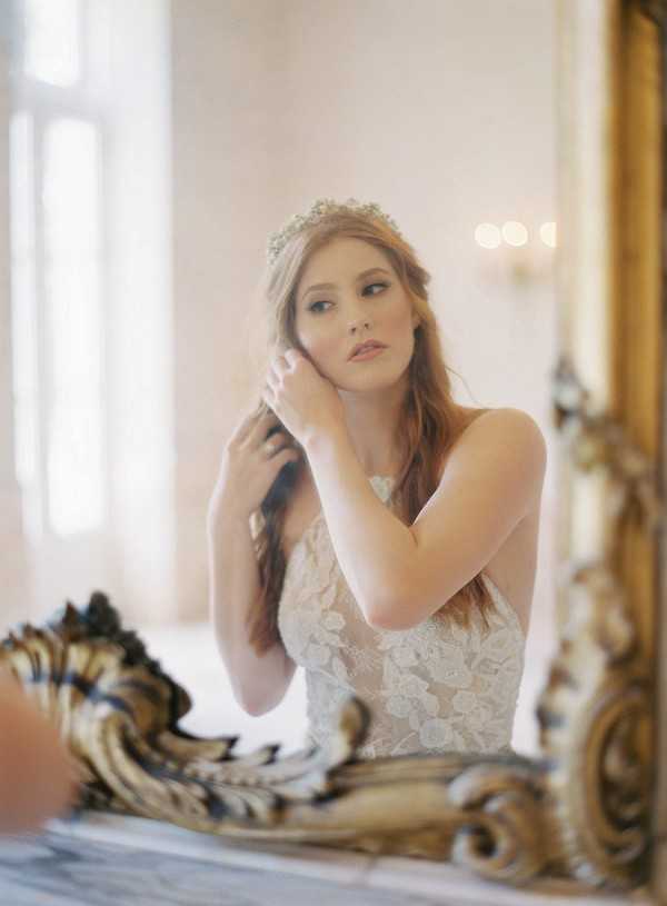 A getting-ready portrait of a bride reflected in a large ornate gold-framed mirror, shot in a softly lit interior room with pale walls and a window visible in the background. The bride wears a sleeveless lace wedding gown and a delicate crystal or pearl bridal crown, and is adjusting her loose auburn hair near her ear. Wall sconces with warm candlelight are visible as soft bokeh in the reflection background. The composition is a close-up portrait framed by the gilded baroque mirror surround, with a shallow depth of field giving the image a soft, airy quality consistent with a classic or romantic editorial styling.