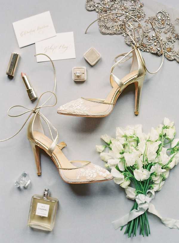 A bridal flat lay arranged on a soft grey surface, centered on a pair of gold stiletto heels with nude mesh toe caps featuring white lace floral appliqué and gold ankle-tie ribbons. Surrounding the shoes are a small bouquet of white sweet peas with grey ribbon, a Chanel No. 5 perfume bottle, a small perfume atomizer, a nude-pink lipstick, a grey velvet ring box containing a diamond engagement ring, two ivory calligraphy place cards with blush pink script, and a heavily crystal-embellished accessory piece in the upper corner. The composition is a styled overhead flat lay with a classic, polished bridal aesthetic.