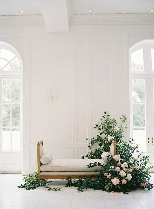 A styled wedding lounge vignette shot indoors in a bright, all-white room with tall arched French doors and white paneled walls with ornate crown molding. A gold-framed daybed upholstered in cream linen sits centered against the wall, flanked by a large asymmetrical floral installation cascading from the right side down to the floor, composed of blush and ivory garden roses, soft pink peonies, and abundant loose greenery with trailing vine stems. The floral arrangement spills organically across the white tile floor, creating a loose, garden-inspired aesthetic. The overall palette is white, cream, gold, blush, and green, with a classic French interior style. Wide shot, no people present. Potential venue feature image.