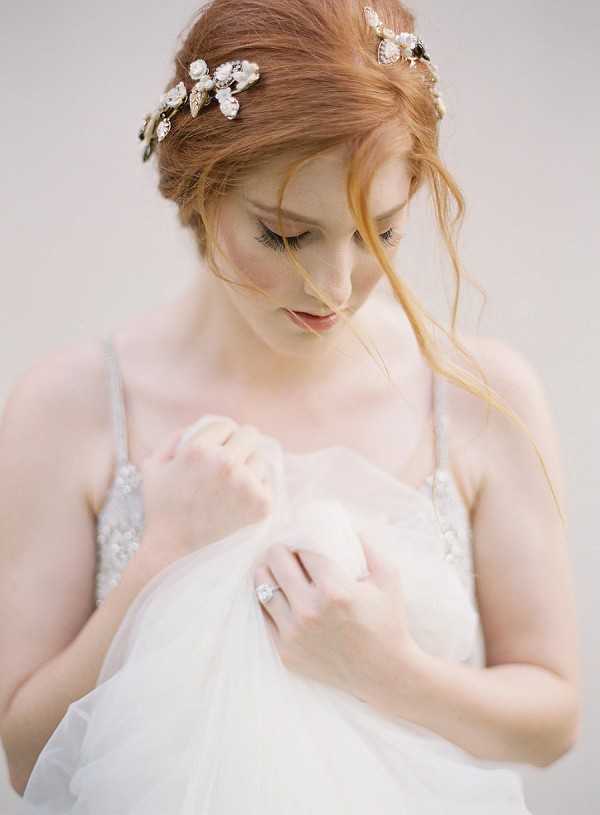 A close-up portrait of a bride with auburn hair styled in a loose updo, adorned with a gold and pearl floral hair vine featuring leaf and flower motifs. She is looking downward and holding soft white tulle fabric in both hands, wearing a white gown with thin spaghetti straps and delicate beaded embellishment. A diamond solitaire engagement ring is visible on her left hand. The shot is tightly cropped from approximately the waist up against a plain, light neutral background, giving it a clean, studio-style aesthetic.