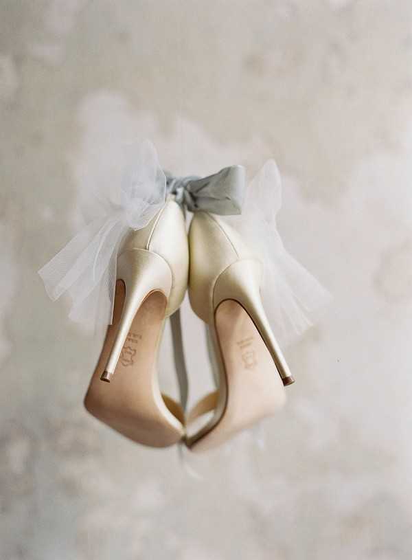 A close-up detail shot of a pair of ivory satin stiletto heels photographed from the back, showing the soles facing the camera. The heels are decorated with a dusty blue-grey fabric bow layered over white tulle, tied at the ankle strap. The soles appear to have handwritten initials or a date inscribed on them, a common bridal personalisation detail. The shoes are displayed against a pale, textured neutral background, and the image has a soft, slightly shallow depth of field with a film-style quality.