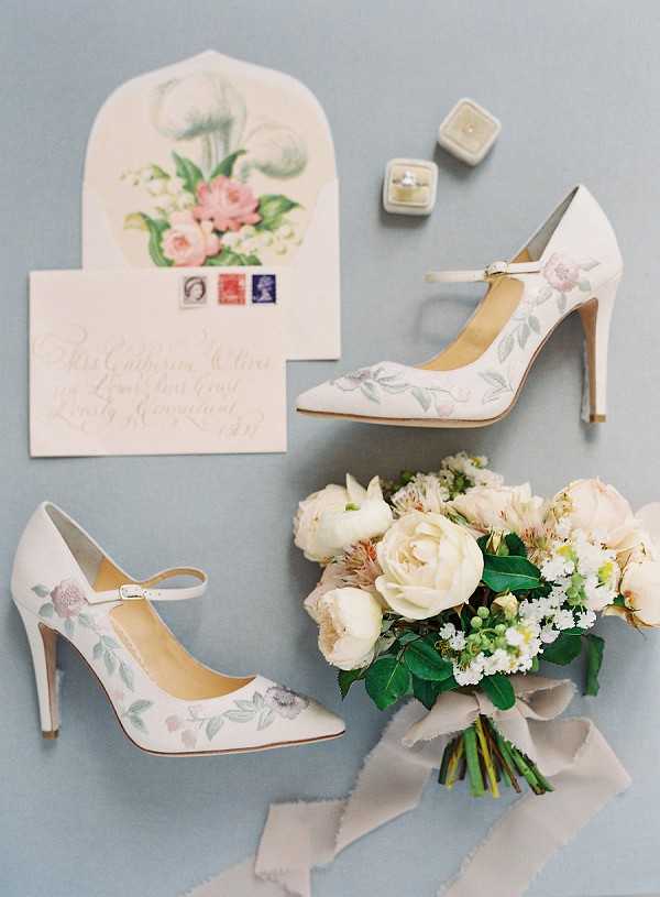 A flat lay detail shot of bridal accessories arranged on a soft blue-grey surface. The centerpiece items include a pair of ivory pointed-toe Mary Jane heels with hand-painted floral and leaf motifs in blush pink and sage green. A bridal bouquet of cream peonies, ranunculus, blush alstroemeria, white waxflower, and green foliage is tied with a long grey silk ribbon. A handwritten calligraphy envelope with a botanical floral illustration inside the flap and UK postage stamps sits alongside a small square velvet ring box in pale grey containing a diamond engagement ring.