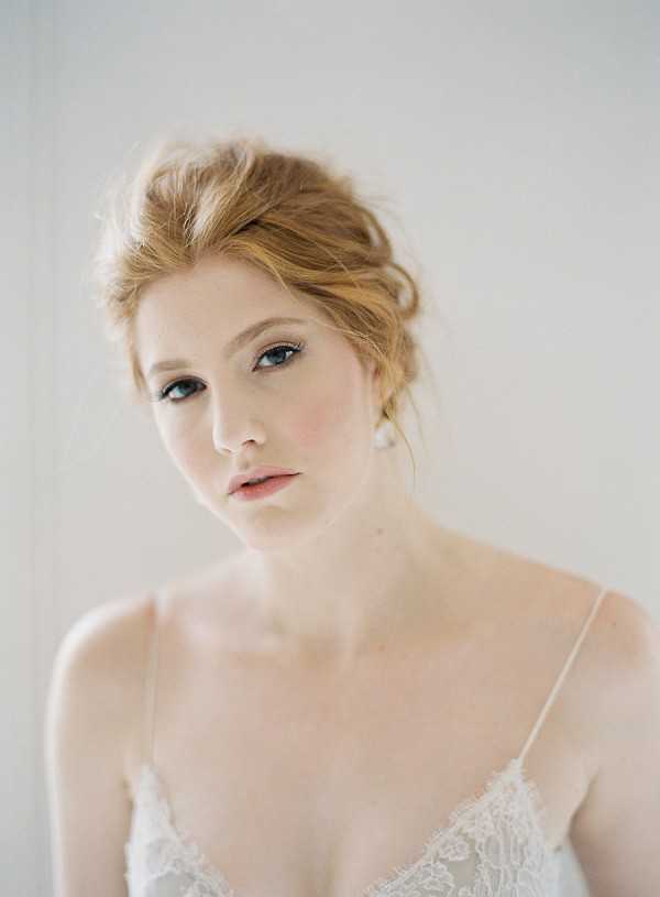 A close-up portrait of a bride during a getting-ready moment, shot against a soft white background indoors. The bride has auburn hair styled in a loose updo with soft tendrils, and wears natural-toned makeup with defined lashes and a soft pink lip. She wears a delicate pearl drop earring and a white lace bridal lingerie or slip with thin spaghetti straps. The composition is a tight head-and-shoulders portrait with shallow depth of field, giving the image a soft, airy quality.