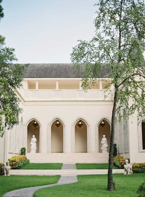 Wide exterior shot of a cream-colored classical building featuring a row of four pointed Gothic-style arched alcoves, each housing a white classical statue, with ornate hanging lanterns visible within the arches. A curved stone pathway leads across a manicured lawn toward a short flight of steps at the entrance. Two additional white cherub or angel statues flank the base of the steps, and ornamental shrubs with yellow and green foliage line the facade. A slender birch tree stands in the foreground. No people are visible in the frame. Potential venue feature image.