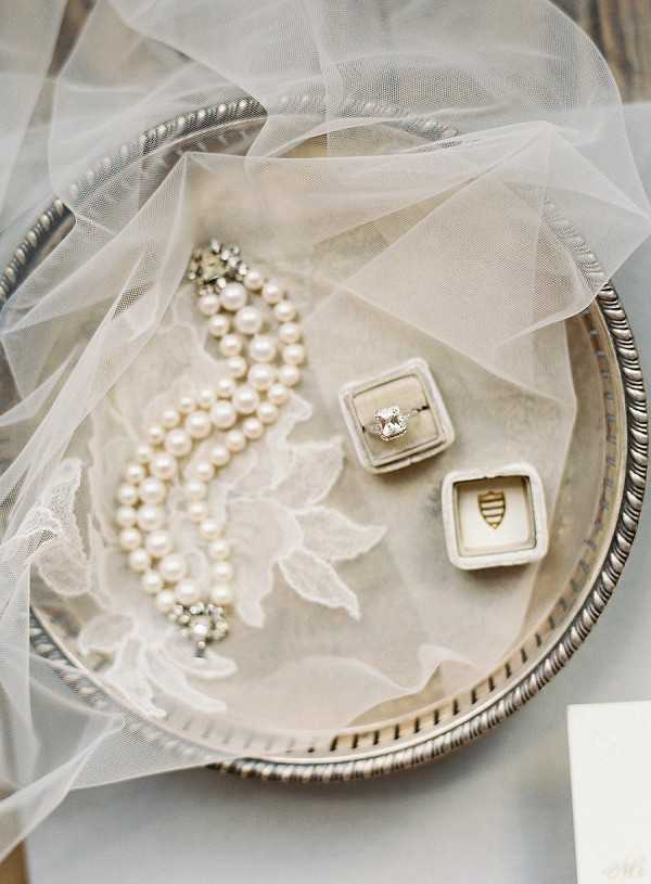 A close-up flat lay detail shot of bridal accessories arranged on a round silver tray with a decorative beaded edge. A double-strand pearl bracelet with crystal rhinestone clasp rests on a sheer tulle veil featuring floral appliqué embroidery in ivory. Two velvet ring boxes in a warm greige tone sit beside the bracelet — one open displaying a cushion-cut diamond engagement ring with a silver band, the other open showing a gold heraldic crest detail. The overall color palette is ivory, cream, and soft silver, consistent with a classic or vintage-inspired bridal styling theme.