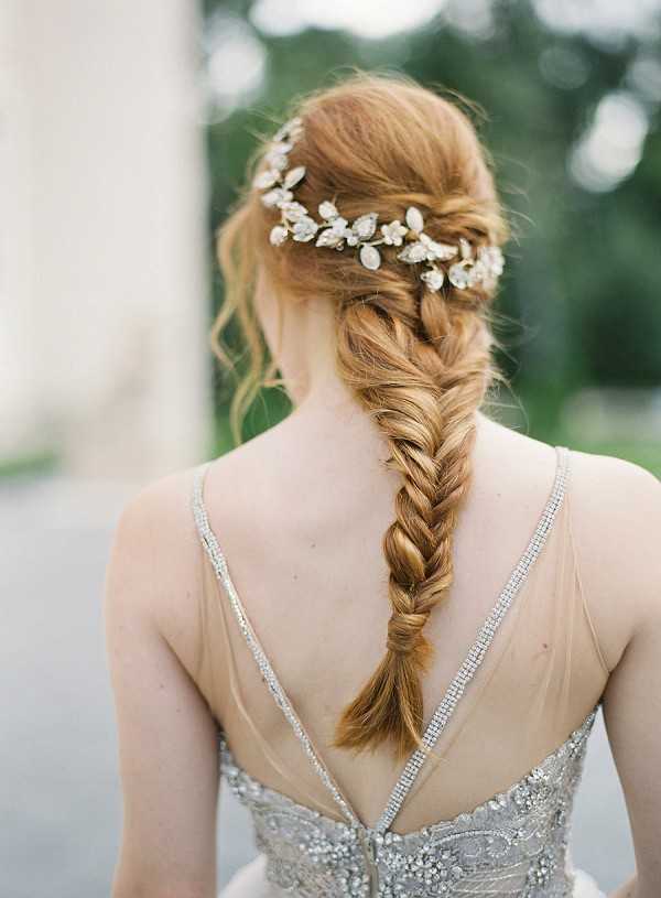 A close-up portrait shot from behind of a bride showcasing her bridal hairstyle and gown back detail. Her auburn hair is styled in a loose fishtail braid running down her back, adorned with a delicate crystal and pearl vine hair piece featuring small floral and leaf motifs in silver. She wears a low-back silver gown with crystal and beaded embellishment along the straps and bodice, with thin rhinestone spaghetti straps forming a deep V at the back. The background is softly blurred with greenery and what appears to be a light-colored architectural column to the left. The overall styling aesthetic is romantic and classical, with fine art film photography characteristics including soft, slightly warm tones.