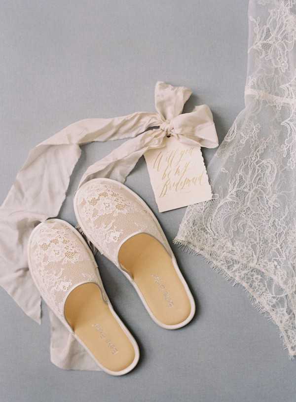 A bridal flat lay detail shot arranged on a muted blue-grey surface. The central items are a pair of ivory lace slip-on shoes with tan leather insoles, accompanied by a cream silk ribbon tied into a bow around a small ivory card with gold calligraphy reading 'Will you be my Bridesmaid.' A length of delicate ivory chantilly lace fabric is draped in the upper right corner of the composition. The overall styling palette is ivory and cream with a soft, minimal aesthetic.