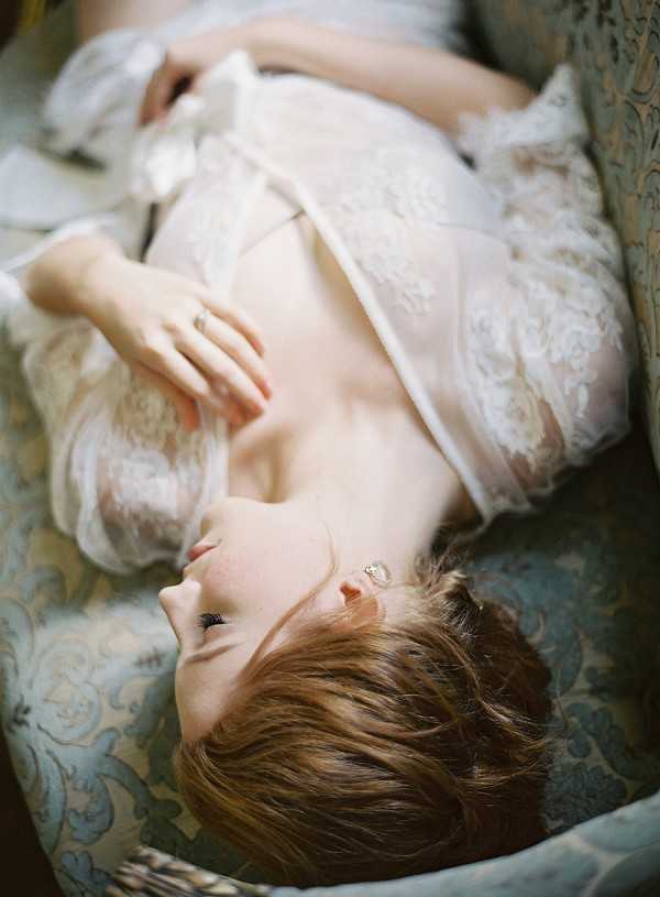 A bride is lying back on an upholstered armchair or chaise with a blue-grey damask fabric pattern, gazing upward in a relaxed pose. She is wearing a white lace bridal garment — likely a robe or lace wedding dress with sheer elements — and a delicate drop earring is visible. Her short auburn hair is loosely styled, and she wears a ring on her right hand, which rests on her chest. The shot is taken from above at a tilted angle, creating a close-up portrait with soft, diffused natural lighting and a romantic, editorial feel consistent with a bridal boudoir or getting-ready session.