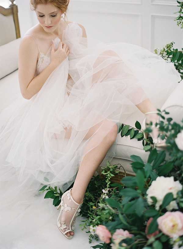 A bridal portrait shot taken indoors in a bright, white-walled room with classic paneling. The bride is seated on a cream upholstered settee, looking downward with one hand resting on her chest. She wears a voluminous ivory tulle ballgown with a lace bodice and sheer layered skirt, paired with nude peep-toe heeled sandals featuring floral lace appliqué detailing. In the foreground, loosely arranged greenery including eucalyptus and leafy branches frames blush pink and white peonies. The composition is a three-quarter portrait with a shallow depth of field that keeps the floral arrangement softly out of focus.