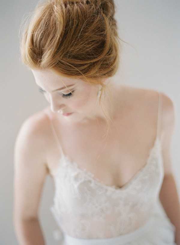 A close-up portrait of a bride shot from slightly above, with soft natural light against a neutral grey-white background. She wears an ivory lace bodice gown with thin spaghetti straps and a V-neckline, featuring delicate floral lace detailing across the chest. Her auburn hair is styled in a loosely pinned updo with soft tendrils framing her face, and she gazes downward with natural makeup including defined lashes. The composition is a tight upper-body portrait with shallow depth of field, giving the image a soft, airy finish consistent with a fine-art getting-ready or bridal portrait style.