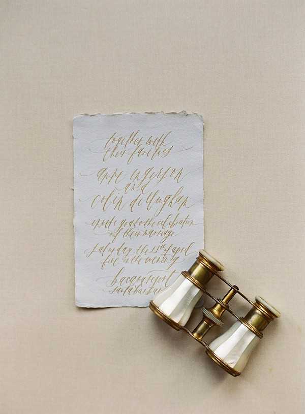 A flat lay detail shot of a wedding invitation written in gold calligraphy script on a piece of white deckle-edged handmade paper, placed on a warm beige linen surface. The invitation text reads 'together with their families' followed by the couple's names and wedding details. A pair of antique brass and mother-of-pearl opera glasses or binoculars is positioned in the lower right corner, partially overlapping the invitation. The overall styling palette is warm gold, ivory, and neutral beige, with a classic and refined stationery aesthetic.