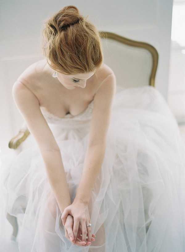 A bride sits on a gold-framed French-style chair in a bright, white interior setting during a getting-ready moment. She is wearing a strapless white tulle ballgown with layers of soft, voluminous skirt, and her auburn hair is loosely pinned up in a relaxed bun. She looks downward with her hands folded in her lap, displaying a solitaire diamond engagement ring, and wears small drop earrings. The image is a close-up portrait taken from a slightly elevated angle, with soft, diffused natural light creating a pale, airy palette throughout the frame.