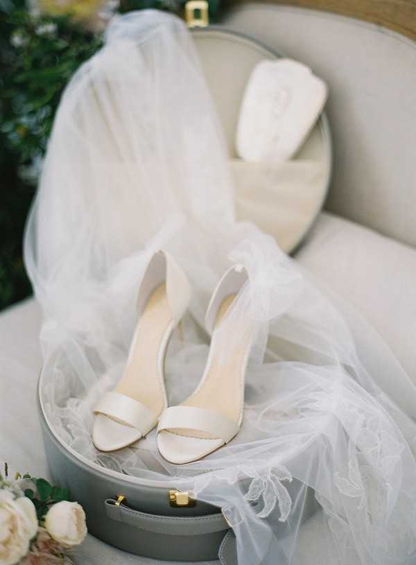 A bridal detail flat-lay shot featuring a pair of ivory satin open-toe heeled sandals placed on top of a grey round hat box with gold hardware. A white tulle veil with lace trim is draped loosely around the shoes and box. A second, smaller round grey hat box sits open in the background, containing a white floral accessory, likely a hair piece. The arrangement is styled on a cream upholstered chair, with cream roses and greenery visible in the lower left corner. The overall palette is ivory, white, and grey with gold accents.