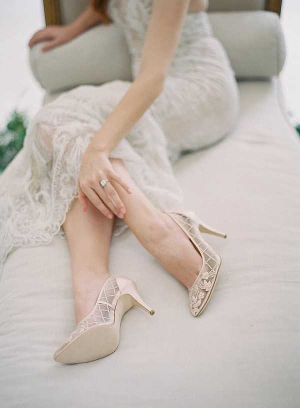 A close-up detail shot of a bride seated on a cream upholstered surface, putting on or adjusting a pair of blush/champagne pointed-toe stiletto heels with lattice cutout detailing and floral appliqué at the toe. The bride is wearing an ivory lace gown with intricate floral lace pattern, and a diamond engagement ring is visible on her left hand. The composition is angled to foreground the shoes while the bride's figure is softly out of focus in the background, creating a shallow depth of field typical of getting-ready bridal photography.