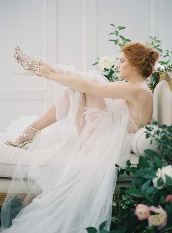A bridal portrait of a single bride seated on a cream upholstered chaise longue or settee against a white paneled wall indoors. She is wearing a layered ivory tulle skirt with a lace bodice and holds up one champagne-gold stiletto heel adorned with floral crystal embellishments, appearing to examine or put it on; she wears a matching heel on the other foot. Her auburn hair is styled in a soft updo, and she wears drop earrings. Surrounding the chaise are lush floral arrangements featuring ivory peonies, blush ranunculus, and trailing greenery. The image is a medium full-length portrait shot with soft, diffused natural lighting and a romantic, editorial styling theme.