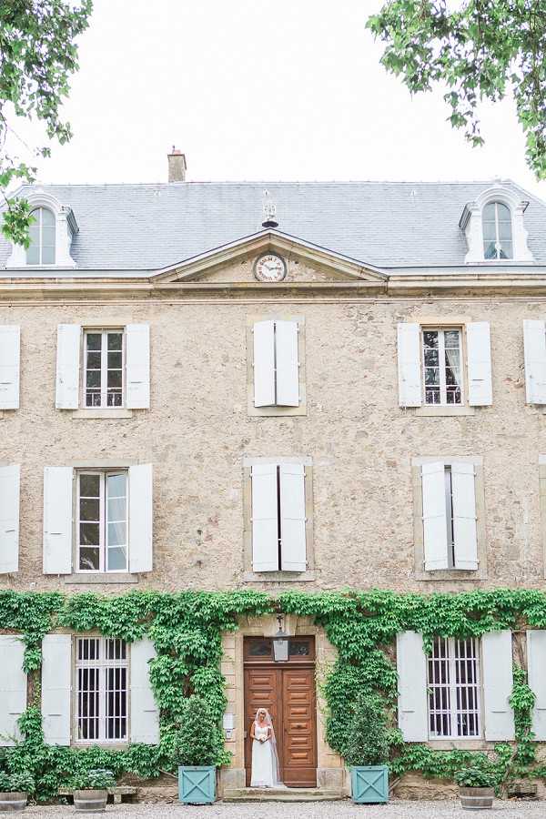 A bride in a white gown and long veil stands in the doorway of a large French chateau, serving as a small focal point against the grand stone facade. The building is a three-story limestone structure with white shutters, a slate mansard roof, a central pediment with a clock, and ivy climbing across the lower level. Teal-painted planters flank the entrance, and the overall decor palette is muted — white, teal, and natural stone. This is a wide exterior portrait shot that primarily showcases the chateau architecture with the bride as a secondary element. Potential venue feature image.