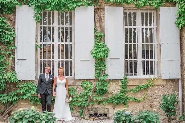 A couple poses together in front of a stone building facade covered in green climbing ivy, with pale grey-blue painted wooden shutters framing tall barred windows. The bride wears a fitted, sleeveless white gown with a V-neckline and holds a small white bouquet, while the groom wears a dark charcoal suit with a light grey tie. The shot is a wide portrait with the couple standing slightly left of center, holding hands and facing the camera. The setting appears to be the exterior courtyard or grounds of a French chateau or mas.