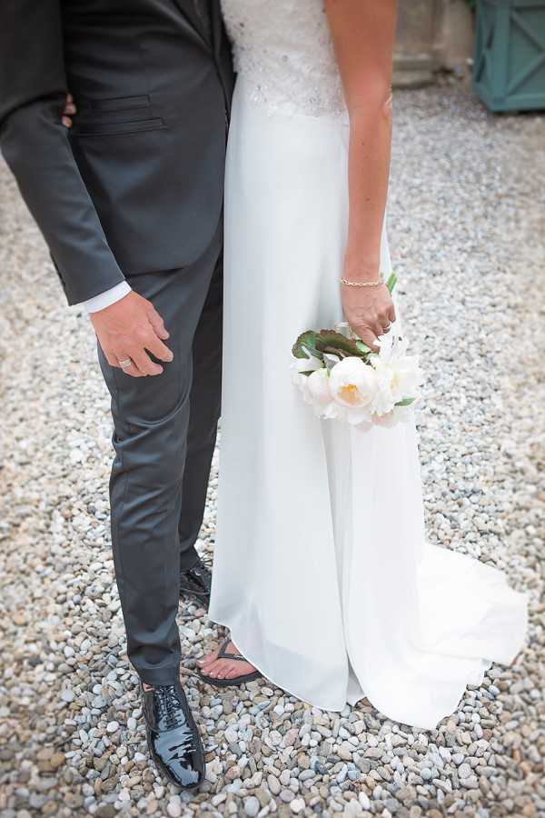 A close-up detail shot of a bride and groom standing side by side on a gravel surface, cropped from the waist down. The bride wears a white gown with a lace-embellished bodice and a flowing skirt with a short train, and holds a small bouquet of cream and blush peonies with green foliage; a delicate gold bracelet is visible on her wrist. She is wearing flat sandals, while the groom wears a charcoal suit with black patent leather Oxford shoes. A wedding band is visible on the groom's hand, and a teal-painted door or structure is partially visible in the background.