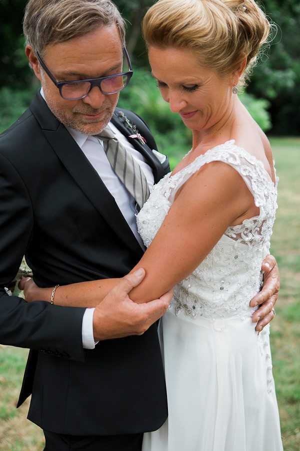 A close-up couples portrait taken outdoors, showing the bride and groom in an intimate moment, both looking downward. The groom wears a dark navy suit with a light grey striped tie, glasses with blue frames, and a small floral boutonniere. The bride wears a white cap-sleeve gown with lace and beaded appliqué detailing on the bodice and a flowing chiffon skirt; her blonde hair is styled in an updo. The groom's hands are gently holding the bride's arm, and she wears a ring visible on her hand. The setting appears to be an outdoor garden or park area, with a natural green background softly out of focus.