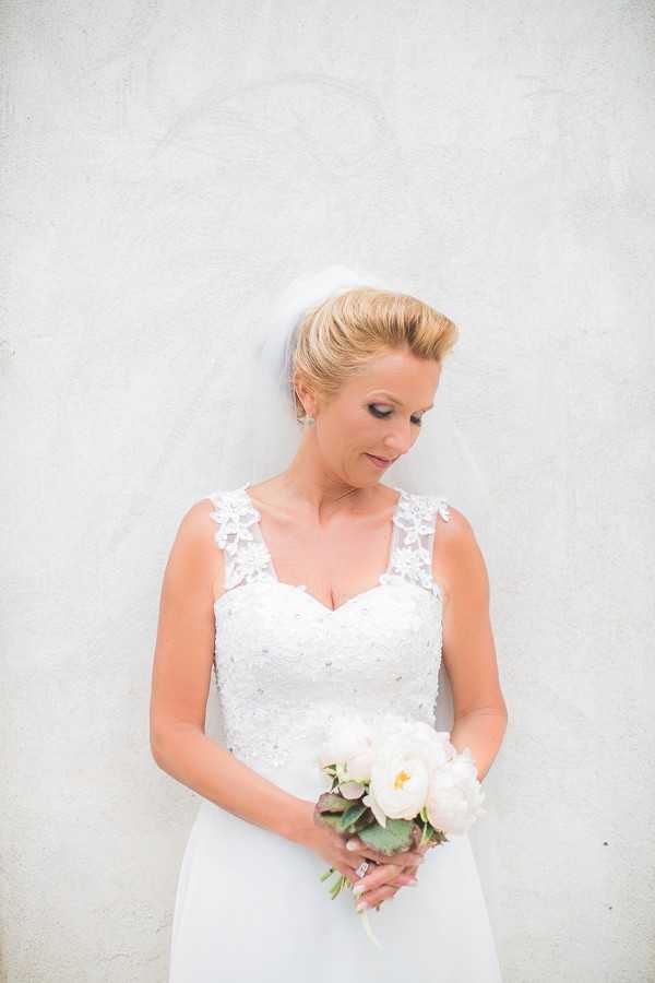 A bridal portrait of a single bride standing against a light-colored rendered wall, looking downward. She wears a white fitted gown with a beaded and embroidered lace bodice, lace strap detailing at the shoulders, and a short veil pinned into an upswept blonde hairstyle. She holds a compact bouquet of white peonies with greenery and looks down toward it. Her accessories include drop earrings and a diamond ring. The image is a medium close-up portrait with a classic, clean styling approach.