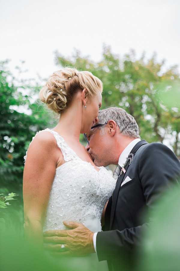 A couple shares a kiss during an outdoor portrait session, framed by foreground foliage in a shallow-depth-of-field composition. The bride wears a white beaded and embroidered sleeveless gown with a V-neckline, and her blonde hair is styled in an updo accessorized with small pearl pins. The groom wears a dark charcoal suit with a white pocket square and a small floral boutonniere, and he has gray hair and glasses. The shot is a close-up portrait taken from a low angle, with soft natural light and a blurred green garden background.