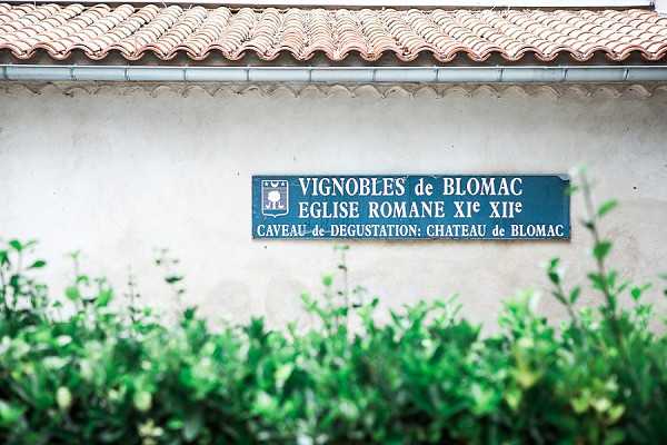 Detail shot of a teal and white directional sign mounted on a rendered white wall beneath a terracotta roof tile overhang, reading 'Vignobles de Blomac – Eglise Romane XIe XIIe – Caveau de Dégustation: Chateau de Blomac.' The foreground features neatly trimmed green hedging. The image establishes the vineyard estate and historic Romanesque church setting of Chateau de Blomac as a wedding venue location. Potential venue feature image.