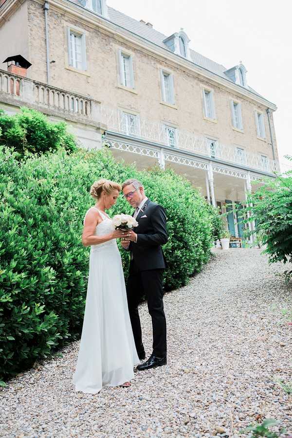 A couple portrait shot outdoors on a gravel path in front of a large French chateau-style building with stone facades, wrought-iron balustrades, and a covered veranda. The bride wears a sleeveless, floor-length white gown with a simple silhouette and holds a compact bouquet of white peonies; the groom is dressed in a dark navy or black suit with a white pocket square. The two are standing close together, heads bowed and touching as they look down at something in their hands, likely exchanging or reading a note. The image is a full-length couple portrait with the chateau prominently visible in the background. Potential venue feature image.