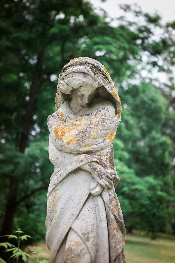 Close-up detail shot of a weathered stone garden statue depicting a draped female figure with her head bowed and arms crossed, set within a wooded garden. The statue shows significant aging with patches of yellow-orange lichen across the grey stone surface. No people are present in the image. The composition is a tight portrait-style shot with a shallow depth of field, leaving the surrounding trees softly out of focus. This appears to be a venue detail or grounds feature shot rather than a wedding moment. Potential venue feature image.