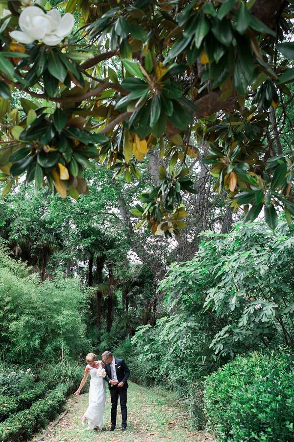 A couple portrait taken outdoors in a densely planted garden, with the bride and groom walking together along a narrow path flanked by low boxwood hedges. The groom wears a dark navy suit and the bride wears a fitted, sleeveless white gown and carries a small white bouquet. The shot is framed from above through the overhanging branches of a magnolia tree, with a single white magnolia bloom visible in the upper left corner, placing the couple small within the frame and emphasizing the lush, layered garden surroundings. The composition is a wide environmental portrait with the couple positioned in the lower third of the image.